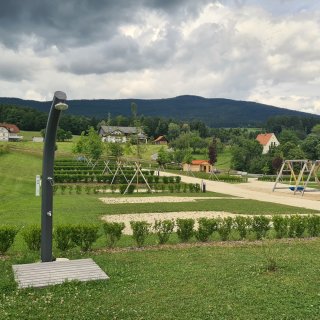 Schwarze Außendusche auf einer Holzplattform
                           mit Spielplatz, parzellierten Flächen, geschotterten Wegen und bewaldetem Bergrücken im Hintergrund.