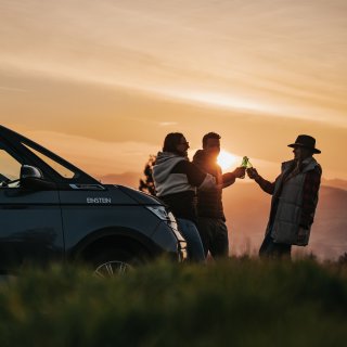 Drei Personen stehen neben einem Camper auf einer Wiese und stoßen mit Getränkeflaschen an. Die Sonne
                           geht hinter den Bergen im Hintergrund unter.