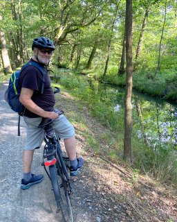 Eine Person mit Fahrradhelm, Sonnenbrille und Rucksack sitzt auf einem schwarzen
                           Fahrrad und blickt in Richtung Kamera. Das Fahrrad steht auf einem unbefestigten Waldweg neben einem schmalen Wasserlauf,
                           der von Gräsern, Sträuchern und Laubbäumen gesäumt wird.