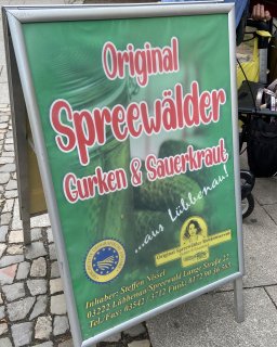 Grünes Verkaufsschild mit der Aufschrift „Original Spreewälder Gurken
                           & Sauerkraut“ auf einem gepflasterten Platz. Auf dem Schild sind außerdem der Hinweis „aus Lübbenau“ und das Zeichen für eine
                           geschützte geografische Angabe zu sehen.