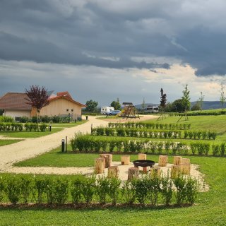 Blick über die begrünte Campinganlage
                           mit parzellierten Flächen, geschotterten Wegen, einer Feuerstelle mit Holzsitzen und einem Spielplatz.