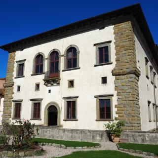 Ein herrschaftliches, zweistöckiges historisches Gebäude mit einer glatten, cremeweißen Fassade unter einem tiefblauen,
                           wolkenlosen Himmel. Die Außenecken des Bauwerks werden von massiven, braungrauen Natursteinquadern stark betont. Die Beletage
                           im ersten Stock zeichnet sich durch drei mittig angeordnete Bogenfenster aus, wobei das mittlere einen kleinen steinernen
                           Balkon mit filigranem Eisengitter besitzt. Ein dunkles Dach mit weitem, hölzernem Überstand schließt das Gebäude nach oben
                           ab. Im Vordergrund grenzt eine niedrige Steinmauer einen gepflegten Gartenbereich mit hellen Kieswegen, exakt geschnittenen
                           Rasenkanten, einem Rosenbusch und einer großen Topfpflanze ab.