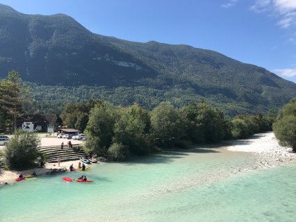 Auf dem Bild ist eine Anlegestellt für Kajaks am türkisblauen
                           Fluss Soča in Slowenien zu sehen. Man sieht einen Parkplatz und Treppen runter zum Wasser und einige Leute auf Kajaks am Flussrand.
                           Im Hintergrund ist das Tal mit bewäldeten Bergen zu sehen.