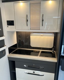 Frontalansicht der Küchenzeile
                           im Hobby Excellent 460 SL mit Arbeitsfläche, Spüle, Kochfeld, Wasserhahn, mehreren Schubladen und Oberschränken. Über der
                           Arbeitsfläche befinden sich Beleuchtung, Steckdosen und ein helles Rollo.
