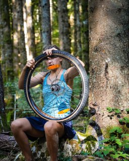 Eine Person sitzt auf einem Stein im Wald
                           und hält ein ausgebautes Fahrradrad in den Händen. Im Hintergrund stehen mehrere Bäume.