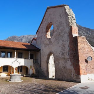 Die alleinstehende,
                           hell verputzte Giebelfassade einer Kirchenruine prägt die rechte Bildhälfte. Das historische Mauerwerk weist ein großes, kreisrundes
                           Loch im oberen Bereich – die Einfassung eines ehemaligen Rosettenfensters – sowie ein hohes, spitzbogiges Portal auf. An den
                           rauen Bruchkanten und einer massiven Stützmauer, an der ein runder Verkehrsspiegel angebracht ist, liegen leuchtend rote Ziegelsteine
                           frei. Links schließt ein zweistöckiges, intaktes Gebäude mit gemauerten Arkadenbögen, einer steinernen Außentreppe und einem
                           traditionellen Terrakottadach an den mit Pflastersteinen versehenen Vorplatz an. Im Hofbereich davor steht ein klassischer,
                           achteckiger Ziehbrunnen aus Stein mit einem filigranen schmiedeeisernen Bogen. Im Hintergrund erheben sich schroffe, graue
                           Berggipfel in einen strahlend blauen, absolut wolkenfreien Himmel.