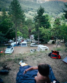 Eine Person sitzt auf einer Isomatte
                           auf einer erhöhten Wiese oberhalb eines Campingplatzes. Unten stehen Zelte, Autos und Wohnwagen zwischen Bäumen. Im Hintergrund
                           sind ein grünes Tal und bewaldete Berghänge zu sehen.