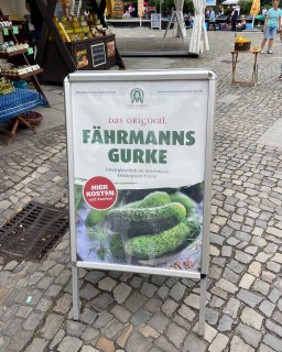 Werbeschild für die „Original Fährmanns-Gurke“ auf einem
                           gepflasterten Platz. Das Schild zeigt mehrere Gewürzgurken und den Hinweis „Hier kosten und kaufen!“. Im Hintergrund stehen
                           Verkaufsstände mit regionalen Produkten.