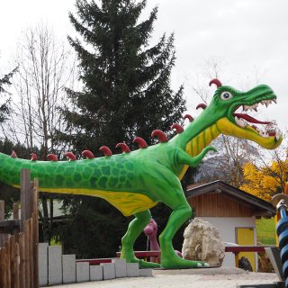 Große grüne Drachenfigur mit rotem Rückenkamm und geöffnetem Maul zwischen
                           Bäumen und Spielgeräten.