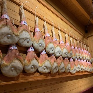 Zahlreiche ganze Schinkenkeulen
                           hängen in zwei dichten, übereinanderliegenden Reihen an einer hellen, horizontal getäfelten Holzwand. Sie sind an Schnüren
                           befestigt und hängen kopfüber herab. Jede Keule ist am Knöchel mit einer auffälligen, weiß-rot gestreiften Banderole markiert,
                           auf der teils die Zahl 