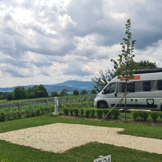 Freie Campingparzelle
                           mit geschotterter Standfläche, Rasen und niedriger Hecke vor Feldern und bewaldeten Hügeln.