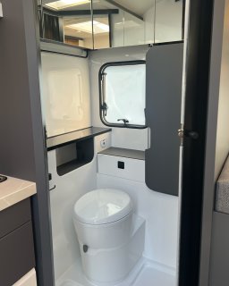 Toilettenbereich im Hobby Excellent
                           460 SL mit weißer Toilette, kleinem Fenster, Ablagefläche, Spiegelschrank und grauer Abtrennung. Links ist ein Teil der Küchenzeile,
                           rechts ein Ausschnitt der Sitzgruppe zu sehen.