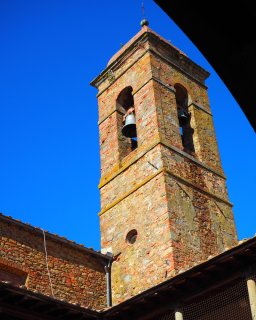 Ein viereckiger Glockenturm aus ungleichmäßig gefärbten, teils fleckigen Back- und Bruchsteinen
                           strebt einem kräftig blauen Himmel entgegen. In der oberen Etage hängen zwei dunkle Glocken gut sichtbar in tiefen Rundbögen.
                           Unterhalb dieser Öffnungen ist ein kleines, kreisrundes Fenster in das Mauerwerk eingelassen. Angrenzend an den Turmschaft
                           sind niedrigere Gebäudeabschnitte mit traditionellen Terrakotta-Dachziegeln und einem dunklen Regenfallrohr zu erkennen. Ein
                           Dachüberstand rahmt die Szene am rechten oberen Bildrand mit einem starken Schattenwurf ein.
