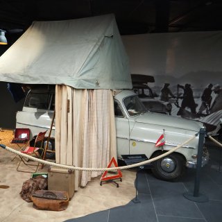 Historischer grauer Pkw mit großem Dachzelt, Campingstühlen,
                           Schreibmaschine und Reisegepäck in einer Museumsausstellung.