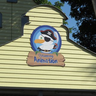 Ein
                           buntes Holzschild an einer gelben Holzwand mit der Aufschrift „Ostseecamp Animation“. Das Schild ist mit einem runden Logo
                           verziert, das eine fröhliche, weiße Möwe im Piraten-Outfit mit Augenklappe, Piratenhut und einem Säbel zeigt.