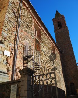 Blick von schräg
                           unten auf die raue Seitenwand einer Kirche, die aus unregelmäßigen Natursteinen und rötlichen Ziegeln gemauert ist. Im Vordergrund
                           steht ein filigran verziertes Eisentor, in das die Jahreszahl 1893 eingearbeitet ist. An der linken Gebäudehälfte führt eine
                           freistehende eiserne Wendeltreppe zu einer einfachen Holztür im Obergeschoss hinauf. Rechts im Bild ragt der dazugehörige
                           kantige Glockenturm mit einem spitz zulaufenden Dach steil in den wolkenlosen, tiefblauen Himmel auf.