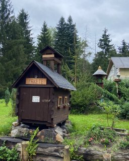 Dunkelbrauner, hausförmiger Holzkasten mit der
                           Aufschrift „Pakete“ in einem begrünten Garten. Der Kasten besitzt eine Tür, zwei kleine Fenster mit Blumenschmuck und einen
                           überdachten Aufbau. Im Hintergrund stehen Nadelbäume, ein Wohnhaus und weitere Gartenelemente.
