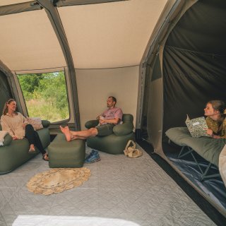 Auf dem Bild sind drei Leute
                           in einem großen Zelt zu sehen, zwei davon sitzen auf Hockern/Couch während eine dritte Person auf einem 