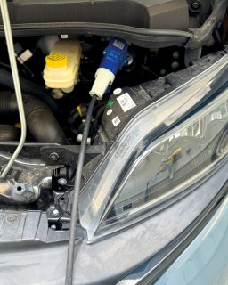 Blick in
                           den Motorraum des Furgok Xpace 600. Deutlich zu sehen ist ein blauer CEE-Stecker, der an einem Kabel hängt und vermutlich
                           für die Stromversorgung oder Ladung genutzt wird. Daneben befindet sich ein Behälter mit gelbem Deckel für Betriebsflüssigkeiten
                           sowie der große Frontscheinwerfer auf der rechten Seite.