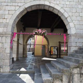 Ein massiver Rundbogen aus hellgrauen, rustikalen Steinquadern bildet den Rahmen des Bildes. Quer
                           durch den Bogen verläuft eine dunkle Stange, die festlich mit pinkfarbenem Band umwickelt ist und von der seitlich lange,
                           pinke Schleifen herabhängen. In der Mitte der Stange ist ein üppiges Gesteck aus rosa und weißen Blumen angebracht. Rechts
                           führen breite, steinerne Stufen in die überdachte Passage. Im schattigen Inneren mit dunkler Holzbalkendecke sind weitere
                           Rundbögen, Informationstafeln und ein steingepflasterter Boden zu sehen, auf den das Sonnenlicht markante Schatten wirft.
                           Im hellen Hintergrund blickt man auf ockergelbe Hausfassaden und ein Schild mit der Aufschrift „Locanda al Municipio“.