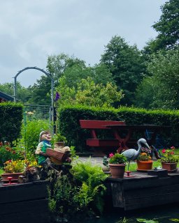 Begrünter Garten am Ufer eines Wasserlaufs
                           mit zahlreichen Blumen, Pflanzgefäßen und mehreren dekorativen Figuren. Im Vordergrund stehen eine Figur mit Schubkarre und
                           eine Kranichfigur; dahinter befinden sich eine rote Sitzgruppe, eine hohe Hecke und Obstbäume.