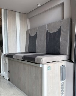 Ein
                           Blick auf eine gemütliche Sitzbank im Wohnbereich des Furgok Xpace. Die Polster fallen durch ein zweifarbiges Design in Hell-
                           und Dunkelgrau auf. Der Unterbau und die angrenzenden Wände sind in einem freundlichen, hellen Holzdekor gehalten.