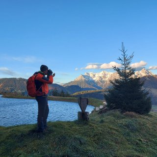 Person mit Rucksack fotografiert an einem Speichersee die verschneite
                           Berglandschaft.