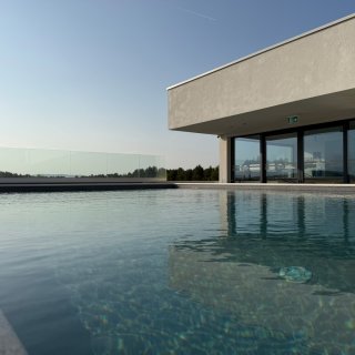 Blick über einen rechteckigen Pool im Camping Resort Šibenik.
                           Am Beckenrand steht ein modernes Gebäude mit großer Glasfront, im Hintergrund sind Bäume und die umliegende Landschaft zu
                           sehen.