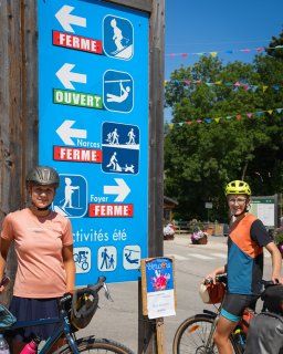 Zwei Personen stehen mit
                           bepackten Fahrrädern neben einem großen blauen Informationsschild mit Symbolen für verschiedene Freizeitaktivitäten. Auf dem
                           Schild sind mehrere Hinweise mit den Aufschriften „FERMÉ“ und „OUVERT“ angebracht.
