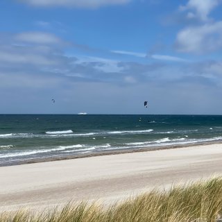 Blick über sanftes Dünengras im Vordergrund auf einen leeren,
                           weißen Sandstrand und die offene Ostsee. Auf den Wellen sind zwei Kitesurfer mit ihren Drachen unterwegs, und ganz am Horizont
                           fährt ein großes, weißes Schiff unter einem leicht bewölkten Himmel.