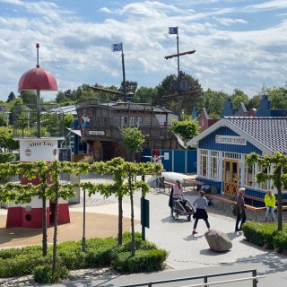 Ein Blick von oben auf den Außenbereich des „Experimentariums“ in Zingst. Auf dem gepflasterten Gelände befinden
                           sich ein rot-weißer Spielzeug-Leuchtturm mit der Aufschrift „Alter Lutz“ sowie ein großes Spielschiff aus Holz mit Piratenflaggen.
                           Ein blaues Holzgebäude mit der Aufschrift „Experimentarium“ steht auf der rechten Seite, während vereinzelte Besucher über
                           den Platz spazieren.
