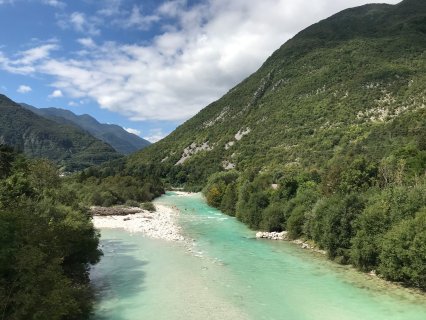 Auf dem Bild ist eine Badestelle am Fluss Soča in Slowenien zu sehen. Der Fluss teilt
                           sich etwas und in der Mitte ist die ein kleiner Steinstrand. Im Wasser sieht man einige Leute und im Hintergrund das weitere
                           Soča-Tal mit Wäldern und Bergen.