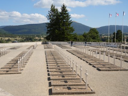 Eine Gedenkstätte für Widerstandskämpfer mit vielen weißen Kreuzen
                           in geordneten Reihen. Rechts stehen mehrere Fahnenmasten mit französischen Flaggen, im Hintergrund sind Bäume, Hügel und ein
                           bewaldeter Berg zu sehen.