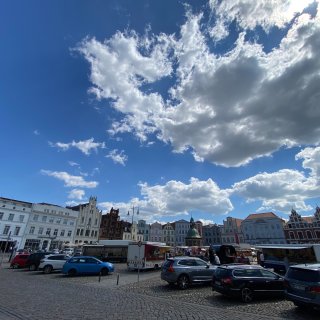 Der weitläufige,
                           kopfsteingepflasterte Marktplatz in Wismar unter einem strahlend blauen Himmel mit großen, weißen Wolkenformationen. Im Hintergrund
                           reihen sich historische Giebelhäuser der Altstadt aneinander, dazwischen ist der Pavillon der Wismarer Wasserkunst zu sehen.
                           Auf dem Platz stehen geparkte Autos und Marktstände.
