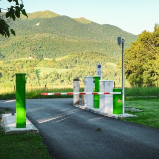 Einfahrt von CAMPING-CAR PARK mit Schranke, Bäumen links und rechts sowie Bergpanorama im Hintergrund.