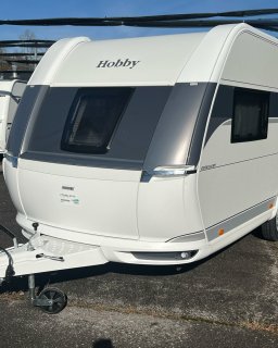Front- und
                           Seitenansicht eines weißen Hobby Excellent 460 SL auf einem asphaltierten Platz. Sichtbar sind die abgerundete Front mit großem
                           dunkel getöntem Fenster, das Hobby-Logo, die Deichsel, ein Seitenfenster und ein Teil der rechten Seitenwand mit grauem Dekorstreifen.