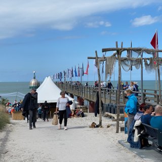 Der belebte Zugang
                           zur Zingster Seebrücke an einem Sandstrand. Im Vordergrund gehen Spaziergänger auf einem befestigten Weg, während rechts Urlauber
                           auf einer Bank sitzen. Die lange Holzbrücke führt weit ins Meer hinein, ist mit zahlreichen bunten Flaggen geschmückt und
                           zeigt am Ende die markante, metallische Tauchgondel Zingst unter blau-weißem Himmel.