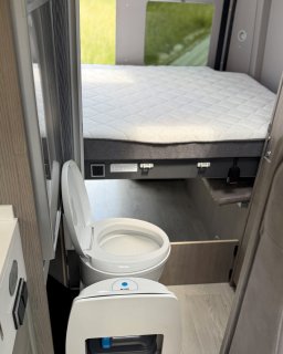 Ansicht der installierten Toilette mit geöffnetem
                           Deckel, positioniert vor dem Heckbett, dessen Matratze sichtbar ist.