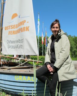 Eine lächelnde Frau in einer hellen wetterfesten Jacke
                           sitzt auf dem Rand eines dekorativen, blau gestrichenen Holzboots auf dem Gelände des Campingparks. Auf dem Segel des Bootes
                           ist das Logo des Platzes mit der Aufschrift „Ostseecamp - Ferienpark, Ostseeheilbad Graal-Müritz“ zu sehen. Im Hintergrund
                           wehen verschiedene Flaggen vor einer Kulisse aus grünen Bäumen unter blauem Himmel.