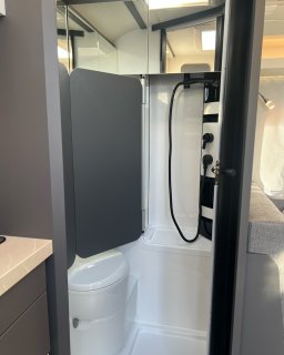 Nasszelle im Hobby Excellent 460 SL mit weißem Toilettenbereich,
                           Duschwanne, Duscharmatur mit schwarzem Schlauch und einer grauen Abtrennung. Darüber befinden sich Spiegelflächen und ein
                           Dachfenster, rechts ist ein Teil der Sitzgruppe zu sehen.