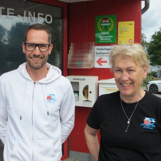 Ein
                           lächelnder Mann im weißen Kapuzenpullover und eine freundlich lachende Frau im schwarzen T-Shirt stehen nebeneinander vor
                           dem roten Eingangsbereich der Rezeption des Ostseecamps Seeblick. Beide tragen das Logo des Campingplatzes auf der Kleidung.
                           Im Hintergrund hängen ADAC- und ACSI-Auszeichnungen an der Wand.