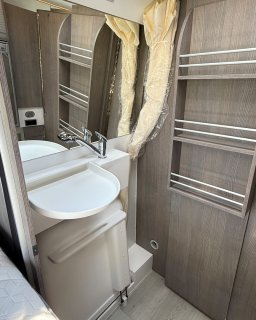 Blick in den kompakten Waschraum des Furgok Xpace 600. Man sieht ein kleines Waschbecken mit Armatur,
                           einen darüber hängenden Spiegel und seitliche offene Regalfächer in Holzoptik. An der Seite ist ein gelblicher Duschvorhang
                           erkennbar.
