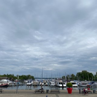 Blick auf den belebten Stadthafen von Waren (Müritz)
                           unter einem wolkenverhangenen Himmel. Am Steg liegen zahlreiche Motorboote, Yachten und Segelboote vor Anker. Im Vordergrund
                           verläuft die gepflasterte Strandpromenade mit Holzbänken, auf denen Passanten sitzen, und einem großen roten Blumentopf.