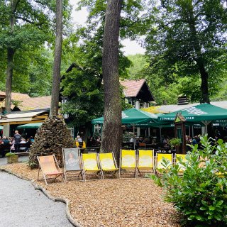 Gastgarten unter hohen Laubbäumen mit zahlreichen
                           besetzten Tischen und grünen Sonnenschirmen. Im Vordergrund stehen mehrere gelbe und braune Liegestühle auf einer mit Holzspänen
                           bedeckten Fläche; dahinter befinden sich dunkel gestrichene Gebäude mit roten Dächern.