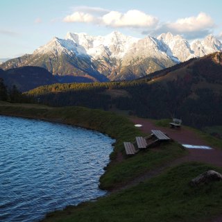 Speichersee mit Sitzbänken vor einem schroffen, teilweise
                           verschneiten Bergmassiv.