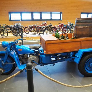Blaues Moto-Guzzi-Lastendreirad mit
                           Holzpritsche vor einer Reihe historischer Motorräder.