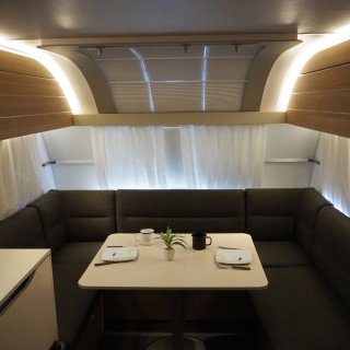 Beleuchteter Esstisch bzw. Essbereich im Inneren des Wohnwagens Adria Adora 593 UP. Man sieht einen kleinen, gedeckten
                           Tisch sowie eine schwarz gepolsterte Sitzecke.