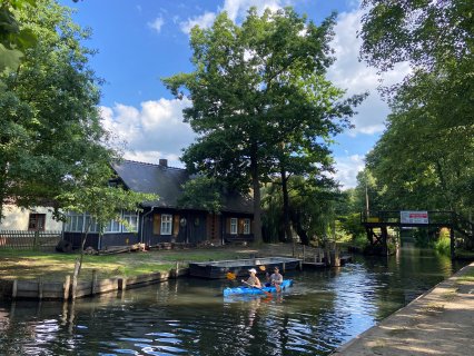 Zwei Personen paddeln
                           in einem blauen Kajak auf einem schmalen Wasserlauf. Am Ufer stehen ein dunkles Holzhaus und mehrere flache Kähne; im Hintergrund
                           führt eine hölzerne Fußgängerbrücke über das von Bäumen gesäumte Wasser.