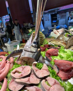Ein reich gedeckter Verkaufsstand auf
                           einem venezianischen Fischmarkt. Im Zentrum thront markant der abgetrennte Kopf eines großen Schwertfisches mit steil aufragendem
                           Schwert. Darum herum präsentieren sich frische, dicke Fischsteaks und Filetstücke auf einem Bett aus hellgrünen Salatblättern
                           und Eis. Im Hintergrund arbeiten Händler in weißen Schürzen unter aufgespannten roten Markisen.