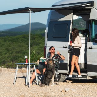 Zwei Personen und ein Hund halten sich neben einem Camper mit ausgefahrener Markise auf. Eine Person sitzt
                           an einem Campingtisch und hält ein Buch in der Hand, während die andere Person in der geöffneten Seitentür des Fahrzeugs steht.